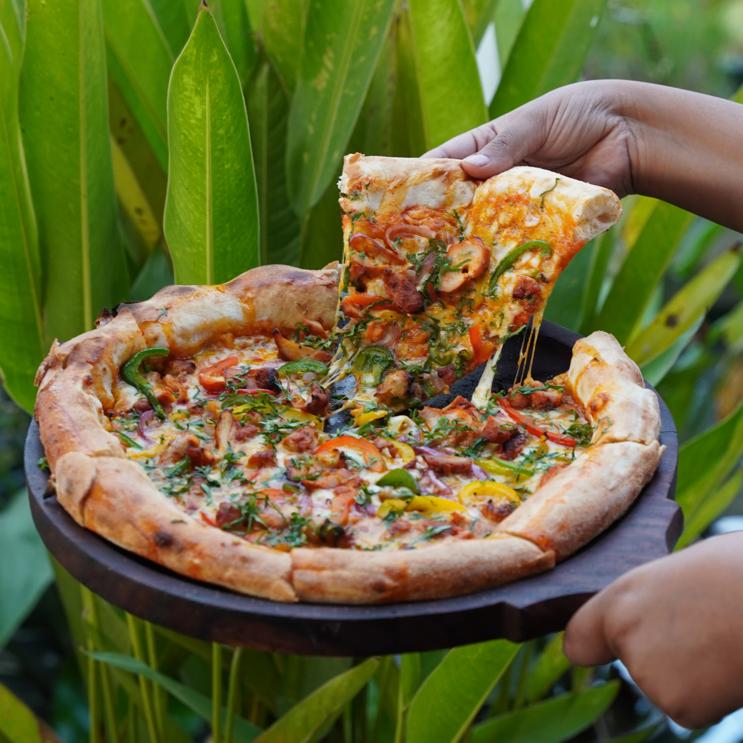 A hand lifting a cheesy slice from a freshly baked pizza loaded with vegetables and toppings at a Bangalore brewhouse.