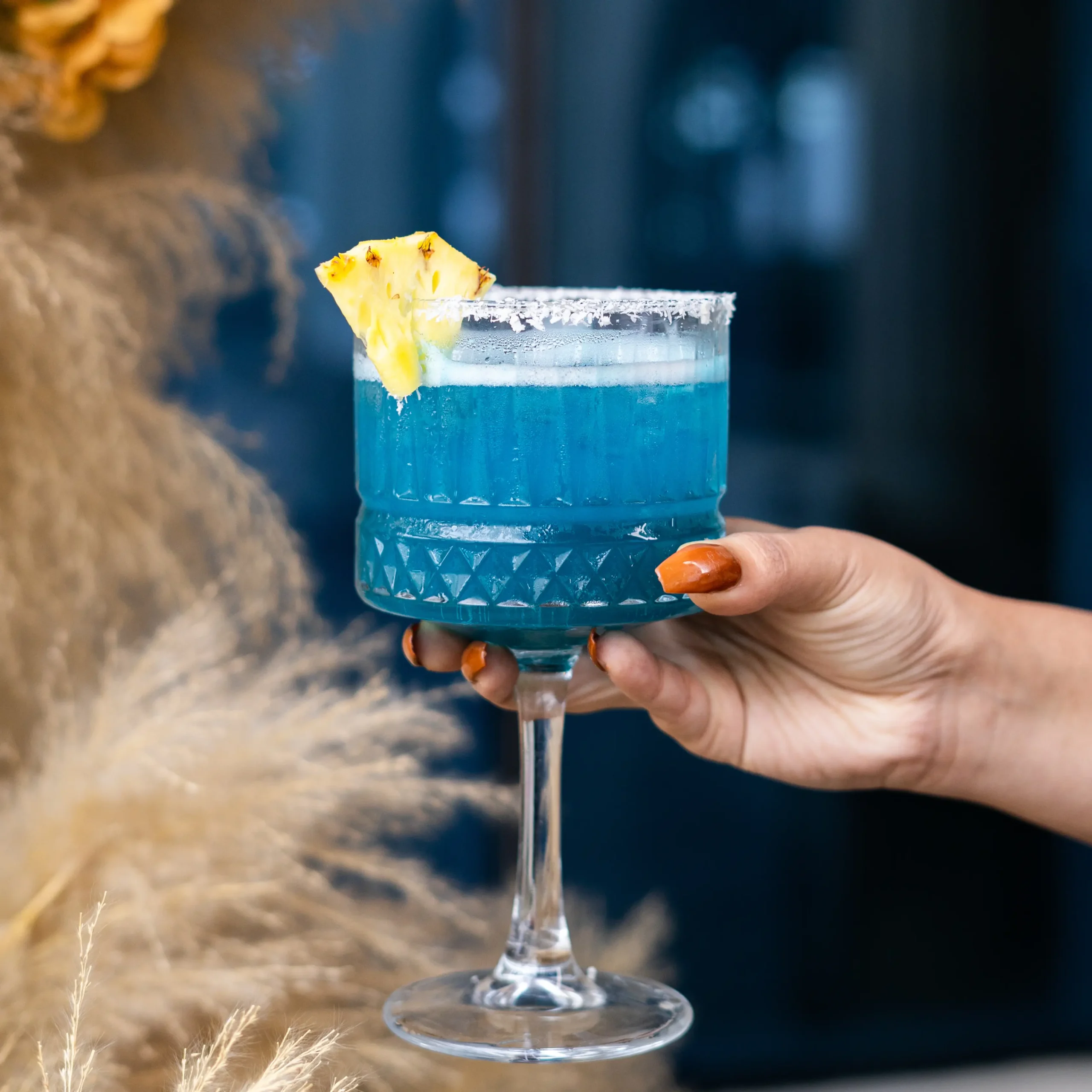 A hand holding a blue tropical cocktail garnished with a pineapple slice at a Bangalore brewhouse.